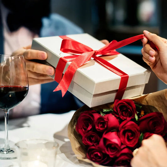 Anniversary gift box with red ribbon, bouquet of red roses, and glass of wine on table