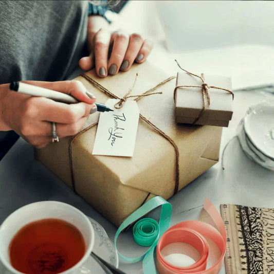 Woman writing thank you tag on wrapped gift box with ribbon, tea, and stationery on table