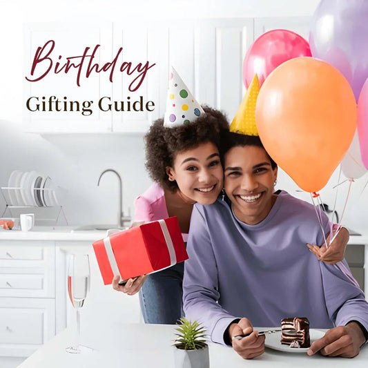 Smiling couple celebrating birthday with cake, balloons, party hats, and gift box in kitchen