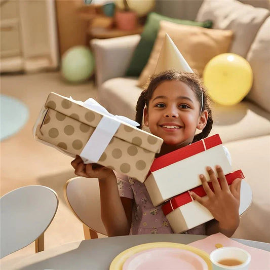 Smiling child with birthday hat holding gift boxes at a party table with decorations