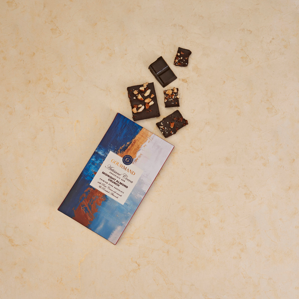 The Gift Studio Gourmand Midnight Almond Crunch chocolate bar with broken pieces on a beige surface.
