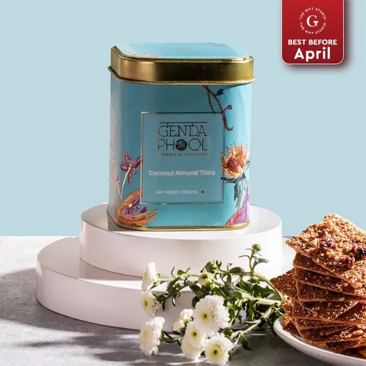 Tin of coconut almond thins with stacked thins on a plate, white flowers in foreground