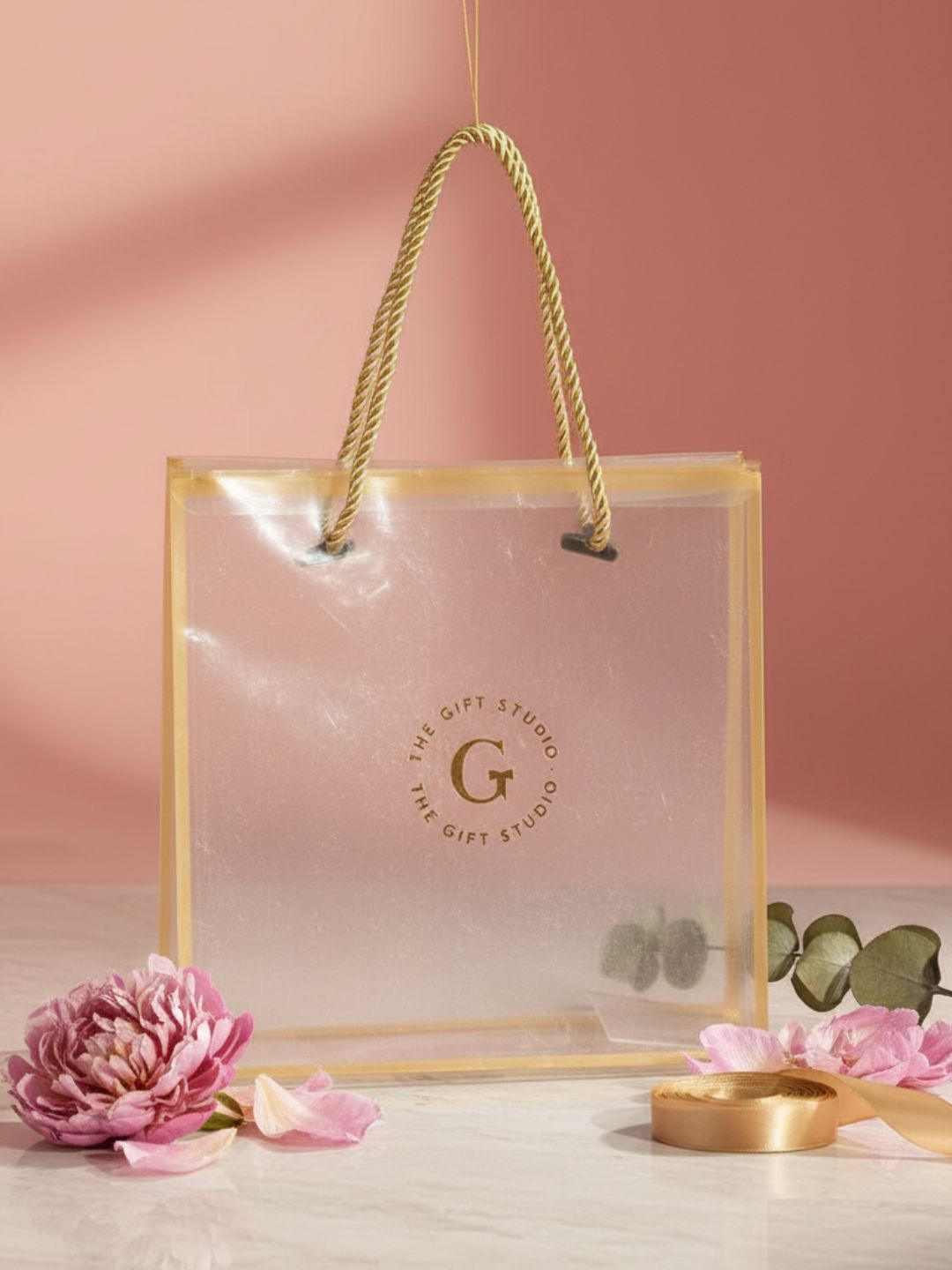 Clear gift bag with gold handle and 'The Gift Studio' logo on a pink background