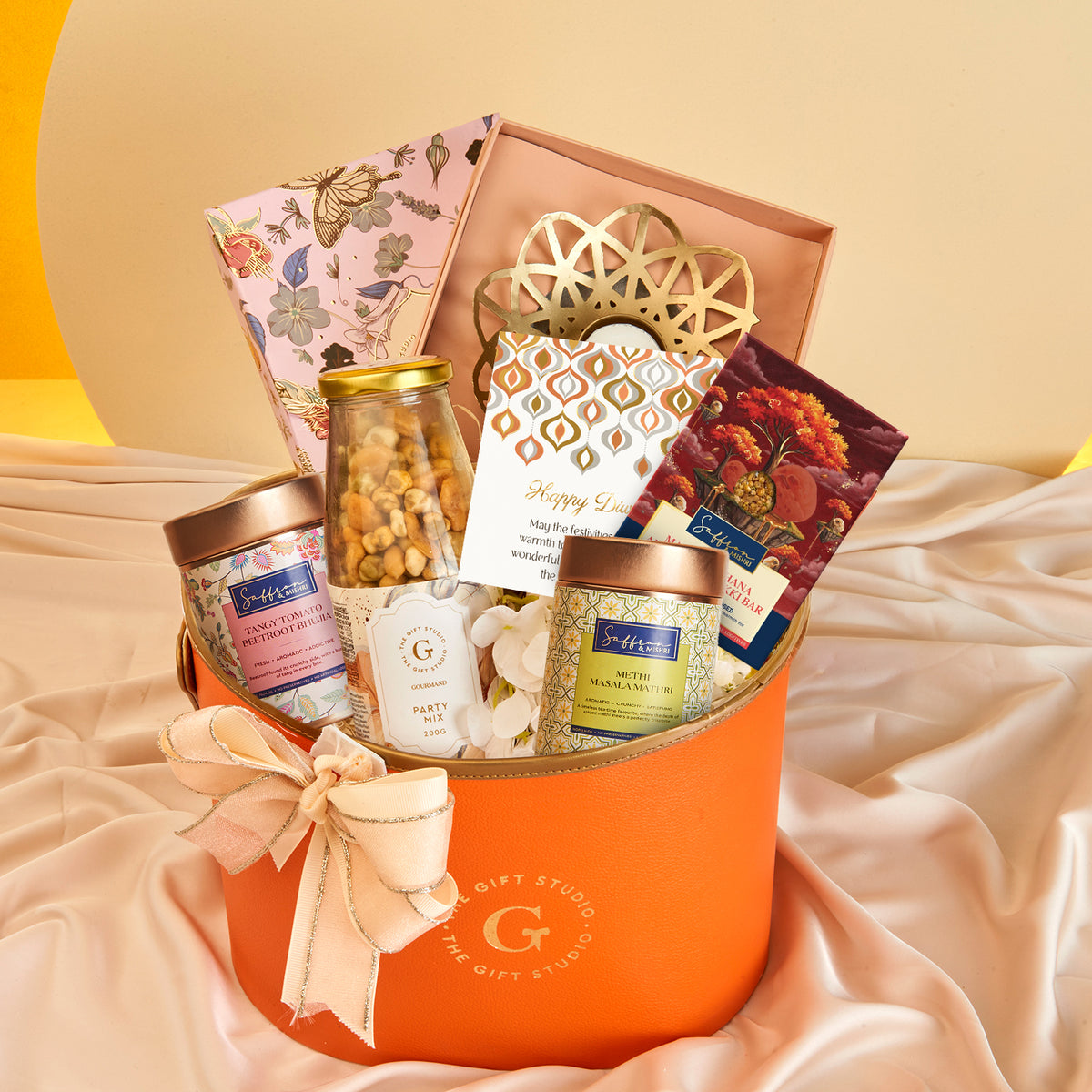 Diwali Floral & Treats Hamper by The Gift Studio featuring gourmet snacks in an orange box.