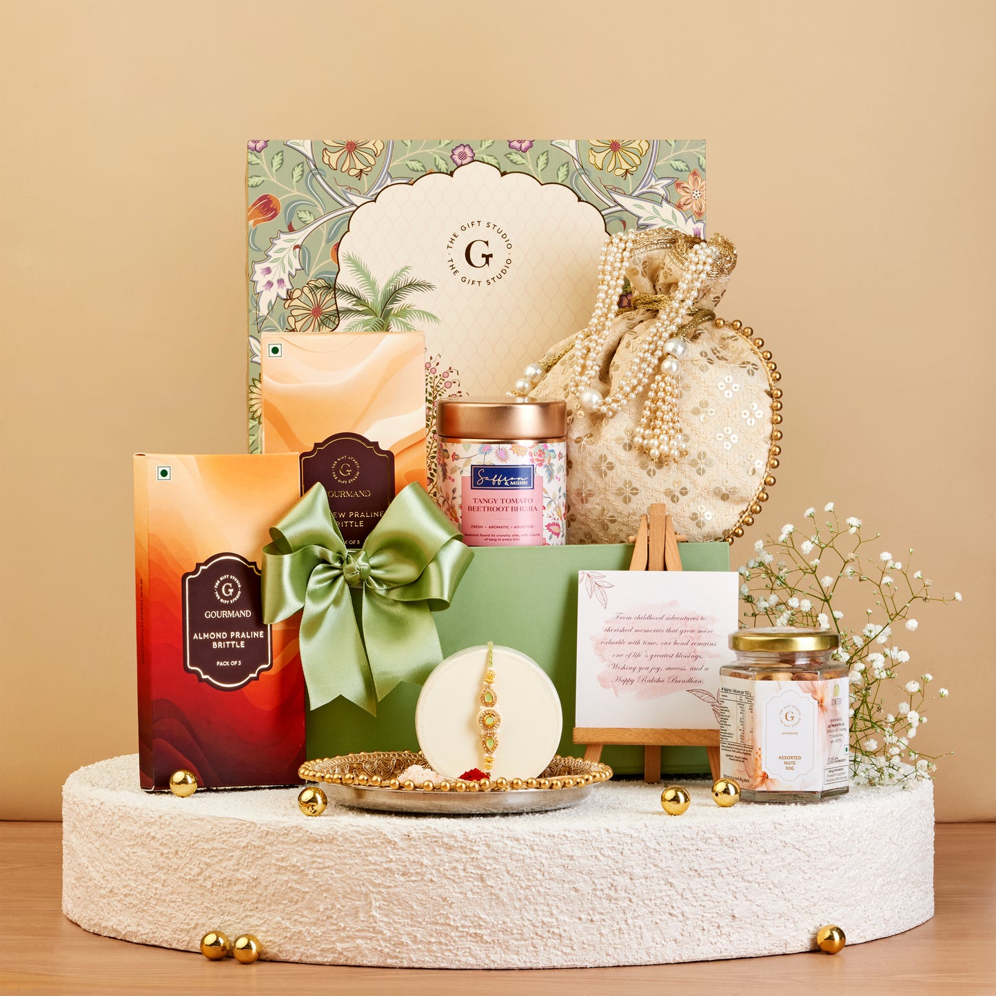 A curated gift set arranged on a round pedestal against a warm beige background. The set includes a large floral gift box, a green ribbon-bow wrapped package, a small orange box, a jar candle, a pearl-beaded pouch, a small framed card, and a glass jar candle, with gold decorative accents.