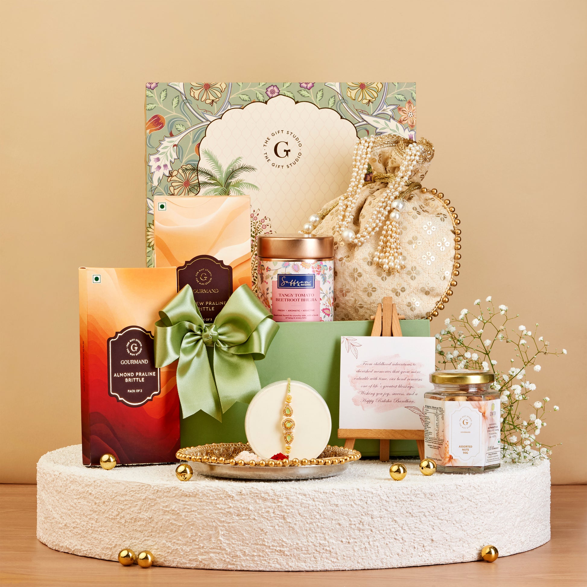 A curated gift set arranged on a round pedestal against a warm beige background. The set includes a large floral gift box, a green ribbon-bow wrapped package, a small orange box, a jar candle, a pearl-beaded pouch, a small framed card, and a glass jar candle, with gold decorative accents.