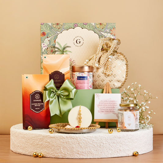 A curated gift set arranged on a round pedestal against a warm beige background. The set includes a large floral gift box, a green ribbon-bow wrapped package, a small orange box, a jar candle, a pearl-beaded pouch, a small framed card, and a glass jar candle, with gold decorative accents.