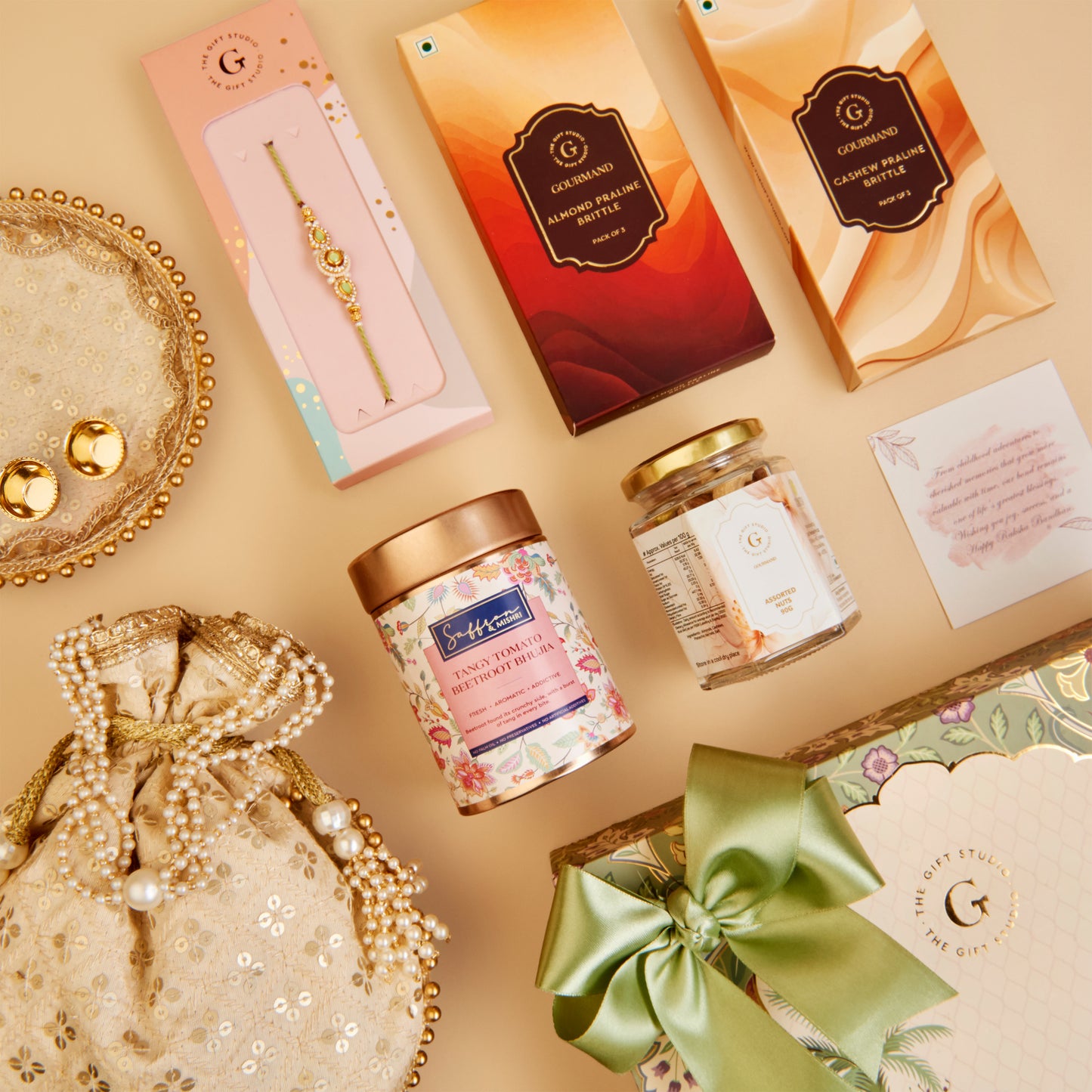 A styled flat-lay of luxury gift items on a warm beige background, including a pink and gold boxed rakhi, two decorative boxes, a floral candle tin, a glass jar, a pearl-trimmed drawstring pouch, and a green ribbon gift box.