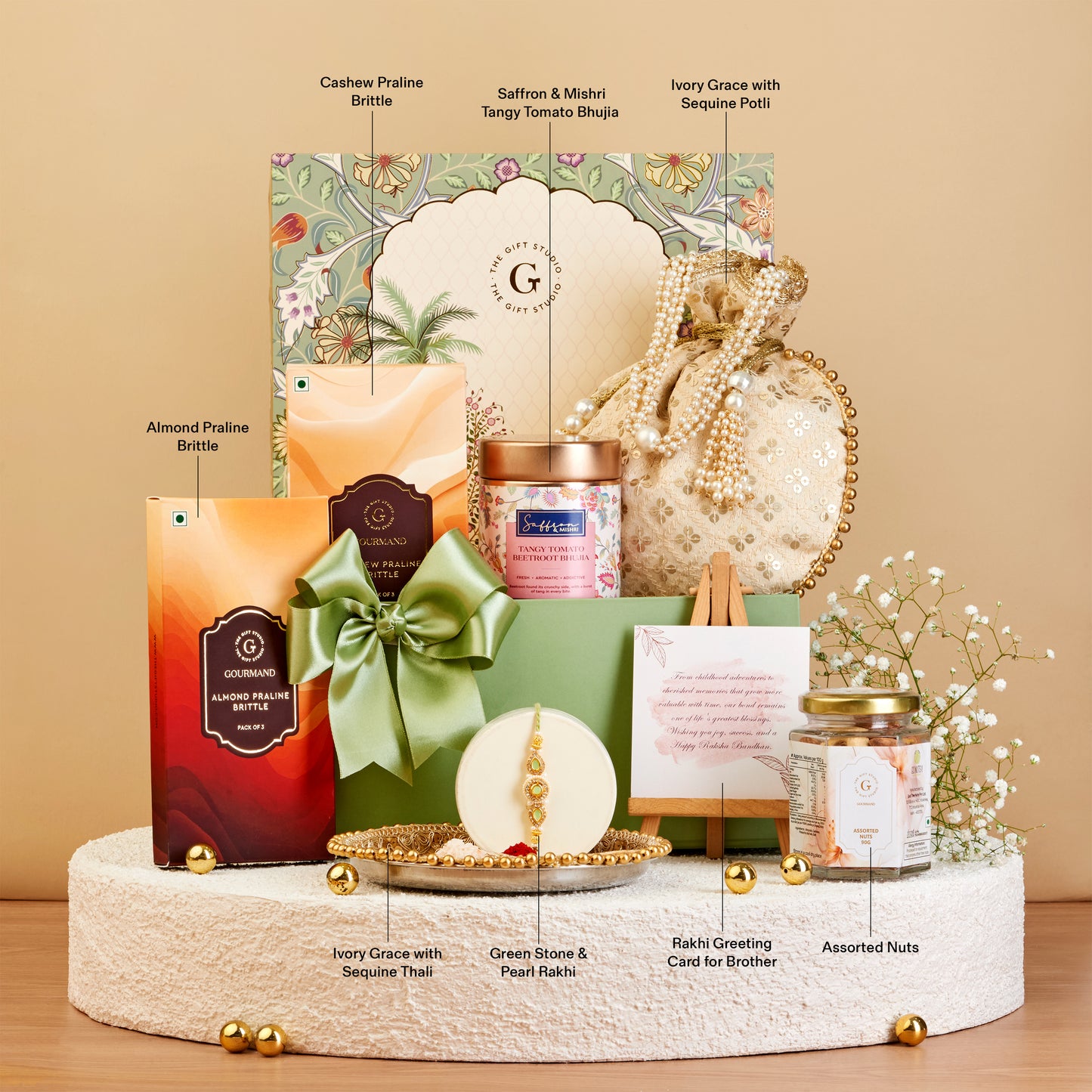 A luxury gift hamper set displayed on a round pedestal against a warm beige background. The set includes a large decorative box with a green bow, a green gift box, a candle, a jar, assorted nuts, and small floral accents.